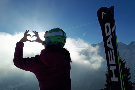 Skifahrerin zeigt Herz in die Luft, rechts ein Ski, dahinter die Berge Skifahrerin zeigt Herz in die Luft, rechts ein Ski, dahinter die Berge