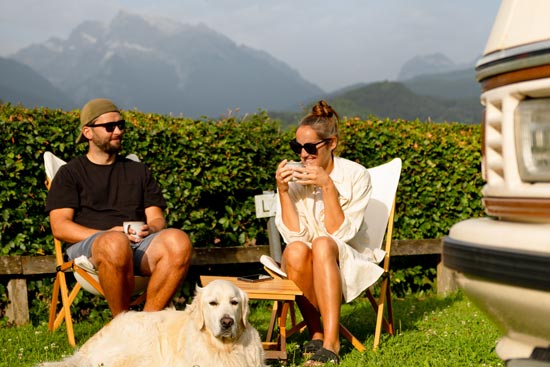 Ein junges Camper-Paar mit Hund, die an einem Sommertag die Aussicht auf die Berge genießen. Ein junges Camper-Paar mit Hund, die an einem Sommertag die Aussicht auf die Berge genießen.